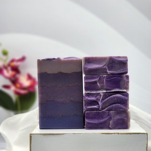 May include: Two bars of handmade soap in shades of purple and lavender. One bar is a tall rectangle with a gradient design and gold accents. The other is a stack of four smaller bars with a marbled pattern. The soaps are displayed on a white wooden stand.