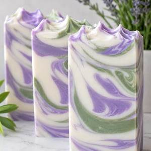 May include: Three rectangular bars of handmade soap with a swirled design. The soap features white, lavender, and green colors. The tops of the bars have a textured, scalloped edge. Lavender sprigs are visible in the background.