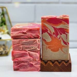 May include: A stack of four pink and red marbled soap bars next to a single bar with orange and red swirls, displayed on a wooden soap dish. The soaps have a textured, layered appearance, suggesting a handmade product.