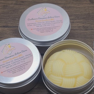 May include: Two silver tins containing solid, yellow, handmade lotion bars. The label on the tin reads "Cashmere Beeswax Lotion Bars" and lists the ingredients: Olive Oil, Shea Butter, Mango Butter, Beeswax, Cashmere Fragrance Oil. The label also states "Net Wt 2.55 OZ/72 g" and includes the company name "Cocoon Creations LLC".