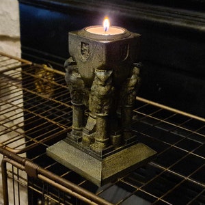 May include: A candle holder made of dark gray stone with a square base and three figures holding up a candle. The candle is lit and the flame is visible.