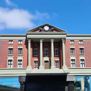 May include: A miniature replica of a brick building with a clock and columns. The building has a red brick facade, white-framed windows, and a grey roof. The clock is centered above the columns.
