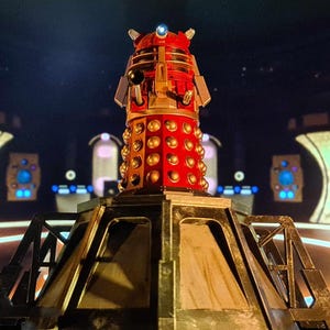 Może przedstawiać: Czerwony i złoty Dalek, fikcyjna pozaziemska rasa z brytyjskiego serialu science fiction Doctor Who, stoi na metalowej podstawie. Dalek posiada kopułę, trzon oka, broń i ramię manipulatora, ze złotymi kulami i akcentami.