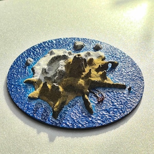 Peut inclure: Un dessous de verre rond et texturé représentant une scène d'île miniature. L'île est représentée avec des nuances de beige, de blanc et de gris, entourée d'un océan bleu profond. De petits rochers gris parsèment le pourtour de l'île.