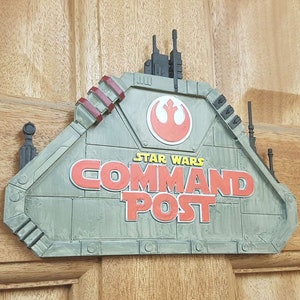 Könnte beinhalten: Ein graues und grünes Star Wars Kommandostand-Schild mit einem rot-weißen Rebellenallianz-Symbol. Das Schild ist aus Kunststoff und hat ein 3D-Design.