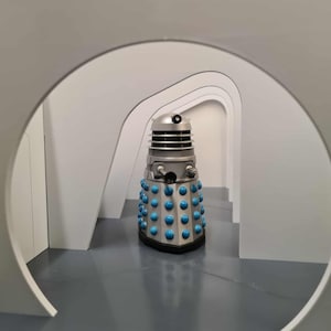 May include: A silver and blue Dalek robot from the Doctor Who series. The robot has a cylindrical body with blue spheres, a silver head with a black eye stalk, and is positioned in a gray hallway.