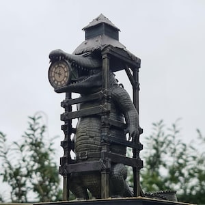 May include: A bronze alligator statue with a clock face in its mouth. The alligator is enclosed in a wooden frame with a small, dark green roof. The clock face has Roman numerals and a decorative design.