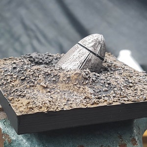 May include: A silver-coloured, metallic, egg-shaped object is partially buried in a brown, textured, square base. The object has a textured surface and a dark line around the middle.
