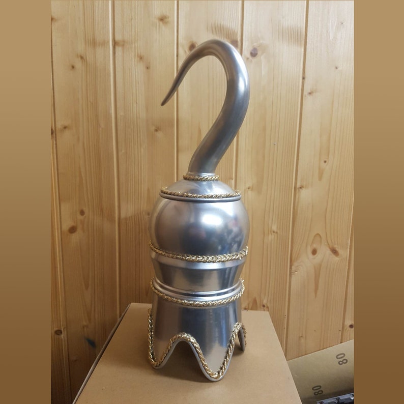 以下が含まれることがあります： 湾曲した刃を持つ銀色の海賊フック。フックは、金色のロープのディテールが付いた丸みを帯びたベースに取り付けられています。ベースには3つの湾曲した足があります。フックは金属製で、小道具または装飾品である可能性があります。