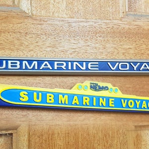 Pode incluir: Dois sinais para o passeio "Finding Nemo Submarine Voyage" na Disneyland. O sinal superior é prateado com letras azuis e o sinal inferior é amarelo, azul e branco com um logotipo do Finding Nemo.