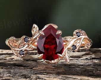 May include: A rose gold engagement ring with a pear-shaped red gemstone surrounded by a delicate vine design with black gemstones.