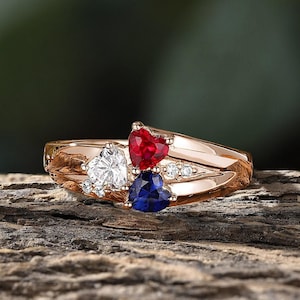 May include: A rose gold ring with three heart-shaped gemstones. The center stone is a white diamond, flanked by a red ruby and a blue sapphire.