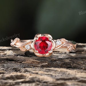 May include: A rose gold ring featuring a round, vibrant red gemstone at its center, surrounded by a floral design. Small, clear accent stones are set around the main stone. The ring's band has a delicate, nature-inspired design.