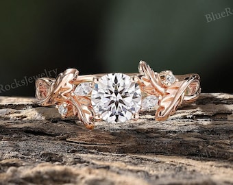 May include: Rose gold engagement ring with a round center stone and leaf accents. The ring is set on a wooden surface.