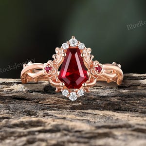 May include: A rose gold ring featuring a central coffin-cut red gemstone, surrounded by small white accent stones and two small red stones on the sides. The band has an intricate vine-like design. The text BlueRocksJewelry appears in the image.