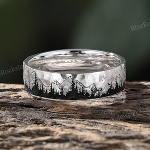 May include: A silver ring with a hammered finish and a black mountain silhouette design. The ring is on a wooden surface.