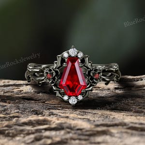May include: A black ring with a large, rectangular, deep red gemstone at the center. The ring features intricate floral and decorative detailing, with smaller red and white accent stones. The ring is set against a natural wood background.