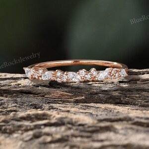 May include: Rose gold delicate marquise and round diamond wedding band.  The band features a unique floral design with pave-set diamonds.  The diamonds are brilliant white and sparkle against the rose gold metal.  The band is thin and dainty, perfect for everyday wear or special occasions.  The image shows the ring resting on a piece of wood.  The watermark 'BlueRocksJewelry' appears multiple times in the image.