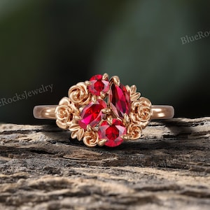 May include: A rose gold ring featuring a cluster of red gemstones. The center stones are surrounded by smaller red gems and rose-shaped accents. The ring is set against a natural wood background.