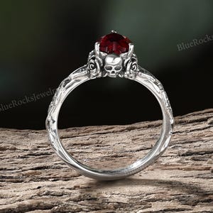 Può includere: Un anello d'argento con una gemma rosso scuro. L'anello presenta un design a teschio e dettagli floreali. La fascia ha un motivo strutturato. L'anello è esposto su un pezzo di legno.