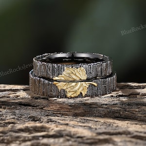 Könnte beinhalten: Zwei Ringe mit einem strukturierten, dunkelgrauen Banddesign. Ein goldfarbenes Blattdetail ziert den oberen Ring. Die Ringe werden auf einem Holzstück präsentiert, mit dem Text "BlueRocksJewelry" im Hintergrund sichtbar.