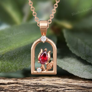May include: Rose gold pendant necklace featuring a red gemstone tulip design. The pendant is set within a rectangular frame and accented with small, sparkling white gemstones. The necklace is displayed on a wooden surface.
