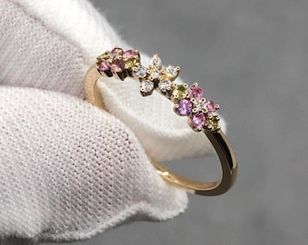 May include: A gold ring featuring a floral design with pink, yellow, and white gemstones. The ring has a delicate band and the gemstones are clustered to resemble flowers. The ring is held by a gloved hand.
