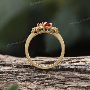 May include: A gold ring with a cluster of red gemstones at the center. The ring has a delicate band and intricate detailing around the gemstones. The background is a blurred view of a wooden surface and greenery. The text "BlueRocksjewelry" is visible.