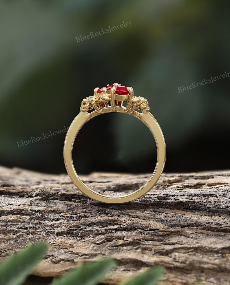May include: A gold ring with a cluster of red gemstones at the center. The ring has a delicate band and intricate detailing around the gemstones. The background is a blurred view of a wooden surface and greenery. The text "BlueRocksjewelry" is visible.