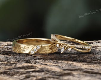 May include: Two gold wedding rings with diamond accents. One ring features an engraved floral design and three diamonds. The other ring has a curved design with multiple diamonds. The rings are displayed on a wooden surface.