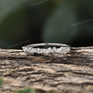 May include: A delicate silver band with a row of small, sparkling diamonds set in a leaf-like pattern. The ring is on a wooden surface.