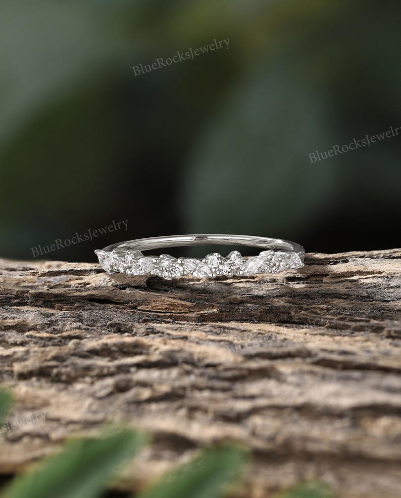 May include: A delicate silver band with a row of small, sparkling diamonds set in a leaf-like pattern. The ring is on a wooden surface.