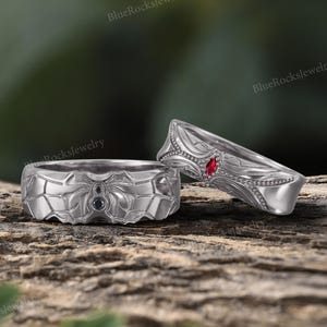 May include: Two silver rings. One ring features a spiderweb design with a spider in the center and two black stones. The other ring has a leaf design with a red stone and small clear stones. The rings are on a wooden surface.