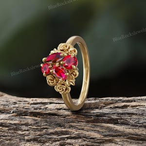 May include: A gold ring featuring a cluster of red gemstones and rose accents. The ring's band is a simple, polished gold, while the gemstones are set in a floral design. The ring is displayed on a wooden surface.