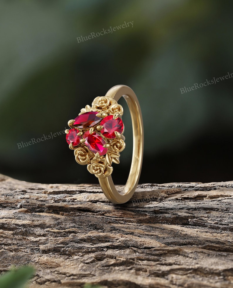 May include: A gold ring featuring a cluster of red gemstones and rose accents. The ring's band is a simple, polished gold, while the gemstones are set in a floral design. The ring is displayed on a wooden surface.