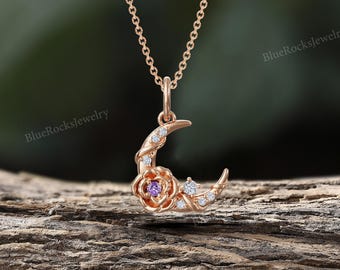 Pode incluir: Colar com pingente de lua crescente em ouro rosa com uma rosa e um detalhe de pedra preciosa roxa. O colar apresenta uma corrente delicada e pequenas pedras preciosas brancas. O fundo é um cenário natural desfocado com o texto "BlueRocksjewelry".