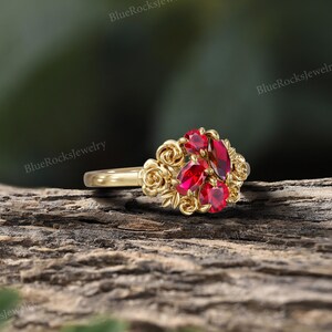 May include: A gold ring featuring a cluster of red gemstones at the center, surrounded by a floral design. The ring's band is smooth and polished, and the gemstones are a vibrant red color. The ring is set against a natural wood background.
