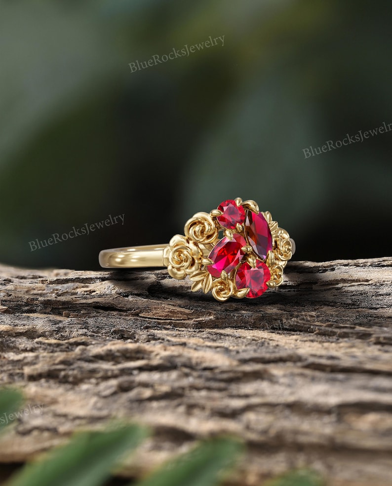 May include: A gold ring featuring a cluster of red gemstones at the center, surrounded by a floral design. The ring's band is smooth and polished, and the gemstones are a vibrant red color. The ring is set against a natural wood background.