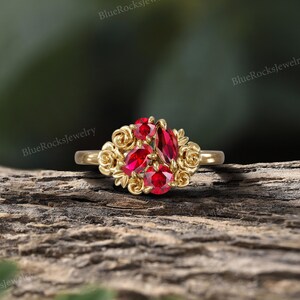 May include: A gold ring featuring a cluster of red gemstones. The gemstones are surrounded by a floral design. The ring is set against a natural wood background. The text "BlueRocksjewelry" is visible in the background.