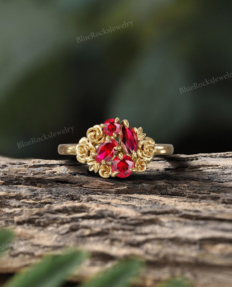 May include: A gold ring featuring a cluster of red gemstones. The gemstones are surrounded by a floral design. The ring is set against a natural wood background. The text "BlueRocksjewelry" is visible in the background.