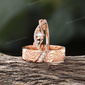 Pode incluir: Um conjunto de an&eacute;is em ouro rosa composto por uma alian&ccedil;a com padr&atilde;o de folhas gravado e tr&ecirc;s pequenas pedras quadradas, e uma alian&ccedil;a curva com uma pedra central de corte marquise rodeada por pequenas pedras redondas e detalhes de folhas.