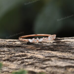 May include: A delicate rose gold band with a row of small, sparkling diamonds set in a leaf-like pattern. The band is shown on a wooden surface.