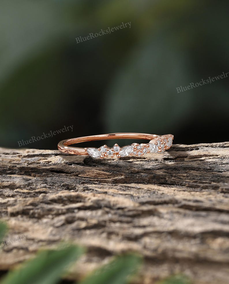 May include: A delicate rose gold band with a row of small, sparkling diamonds set in a leaf-like pattern. The band is shown on a wooden surface.