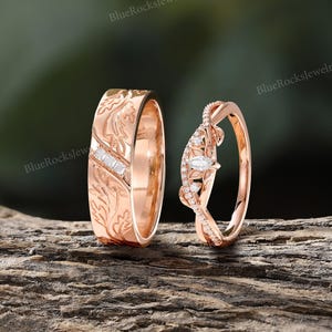 Pode incluir: Um conjunto de alian&ccedil;as em ouro rosa com uma banda larga com padr&otilde;es de folhas gravados e tr&ecirc;s pequenas pedras quadradas, ao lado de uma banda curva com uma pedra central lapida&ccedil;&atilde;o marquise e pequenas pedras redondas, com o texto BlueRocksJewelry.