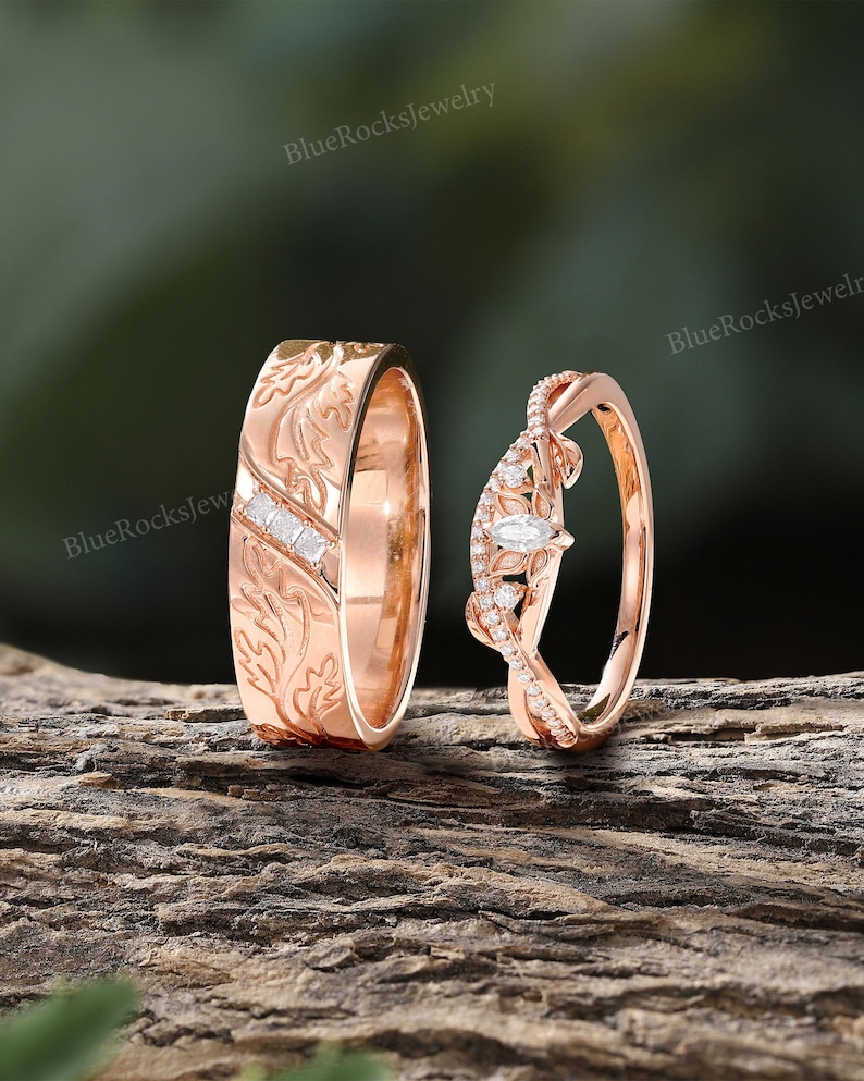 Pode incluir: Um conjunto de alian&ccedil;as em ouro rosa com uma banda larga com padr&otilde;es de folhas gravados e tr&ecirc;s pequenas pedras quadradas, ao lado de uma banda curva com uma pedra central lapida&ccedil;&atilde;o marquise e pequenas pedras redondas, com o texto BlueRocksJewelry.