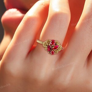 May include: A gold ring featuring a cluster of red gemstones and small gold rose accents. The ring's band is thin and delicate, with the words "BlueRocksJewelry" visible.