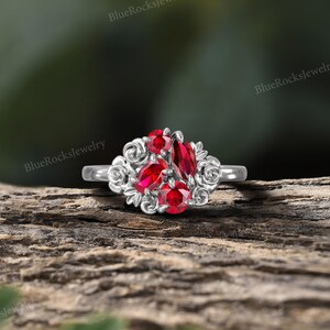 May include: A silver ring featuring a cluster of vibrant red gemstones. The ring's design includes a central marquise-cut stone surrounded by smaller round and pear-shaped gems, accented by floral filigree. The ring is set against a natural wood background.