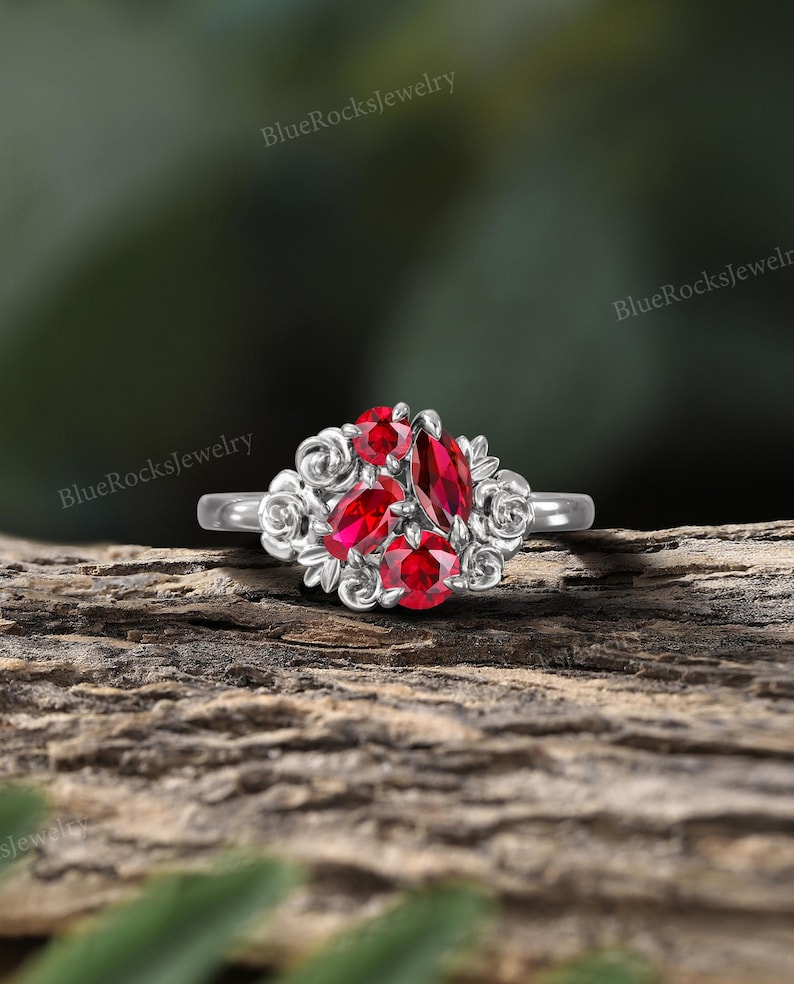May include: A silver ring featuring a cluster of vibrant red gemstones. The ring's design includes a central marquise-cut stone surrounded by smaller round and pear-shaped gems, accented by floral filigree. The ring is set against a natural wood background.