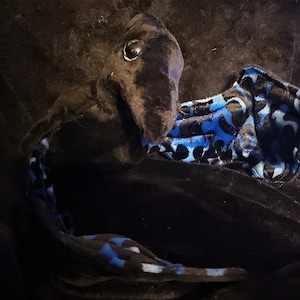 May include: A plush black creature with a long neck and a patterned blue and black belly. The creature has a large, round eye and is made of soft fabric. The overall design is whimsical and playful.
