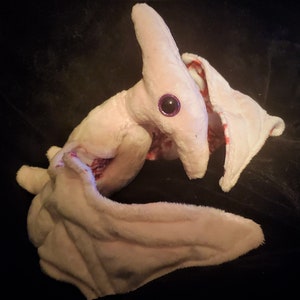 May include: A plush pterodactyl toy in soft pink fabric with large, glittery purple eyes. The toy has a long neck, wings, and a beak-like structure. Red accents are visible on the inside of the wings. The toy is lying on a black surface.